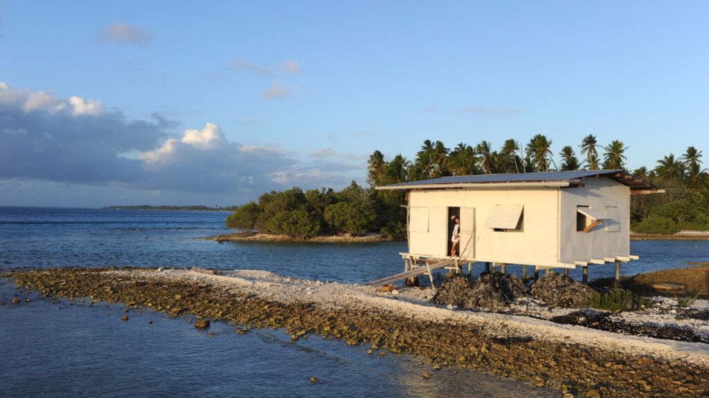 Plywood farm hut on stilts at the reef edge in evening light, a worker in the doorway